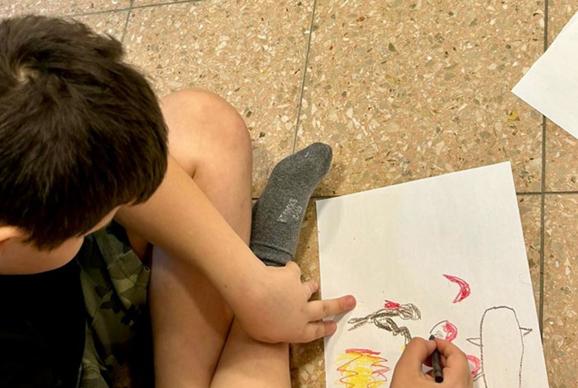 Gutt med mørkt hår sitter på gulvet med beina i kors og tegner et menneske med våpen og ild. Foto: SOS-barnebyer Israel
