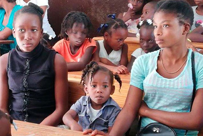To tenåringsjenter og ei lita jente sitter ved en skolepult i ett barnevennlig senter. Foto: SOS-barnebyer Haiti