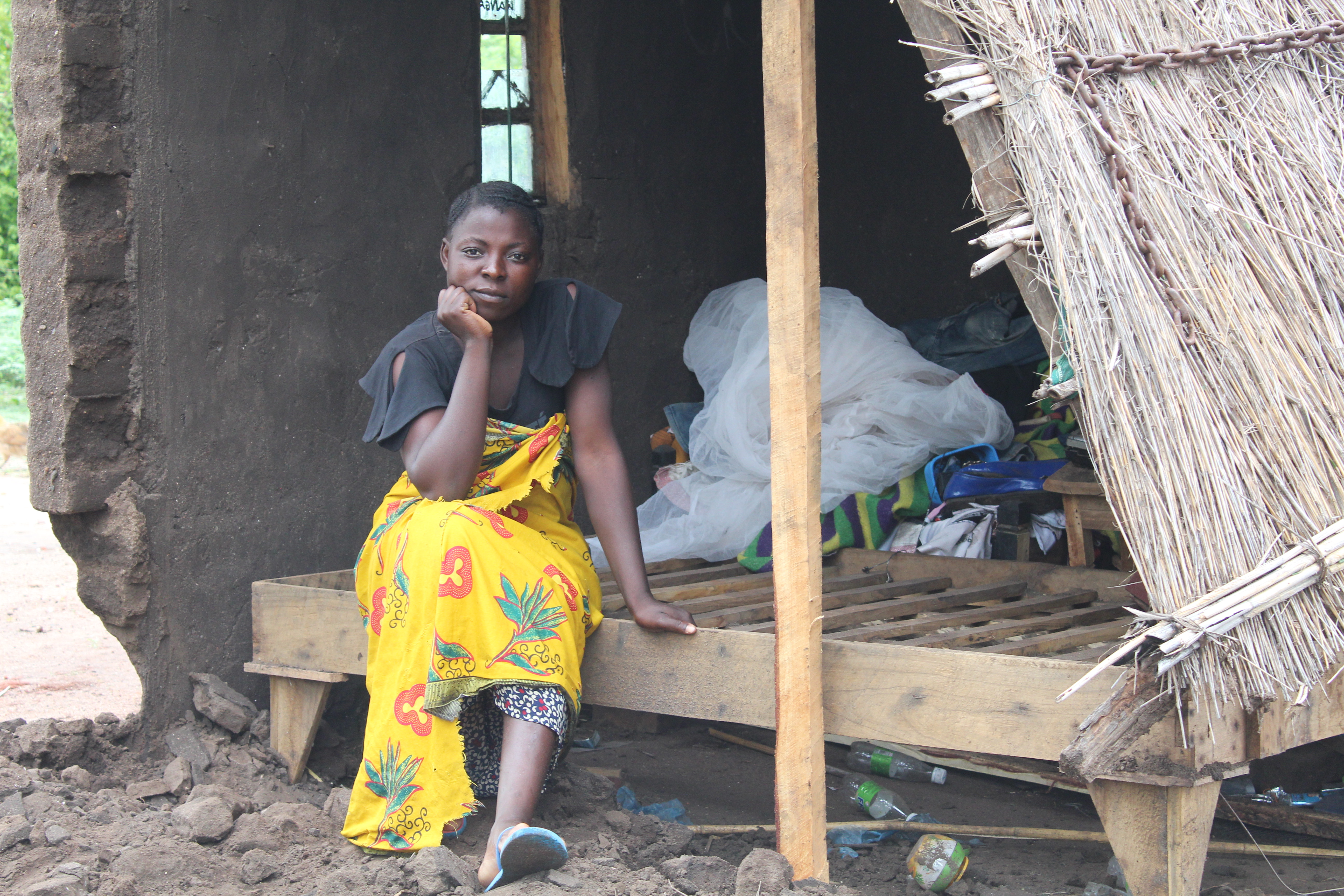 Jenta med kort hår mørk overdel og gult skjørt sitter i et ødelagt hus i Malawi. Foto: SOS-barnebyer Malawi