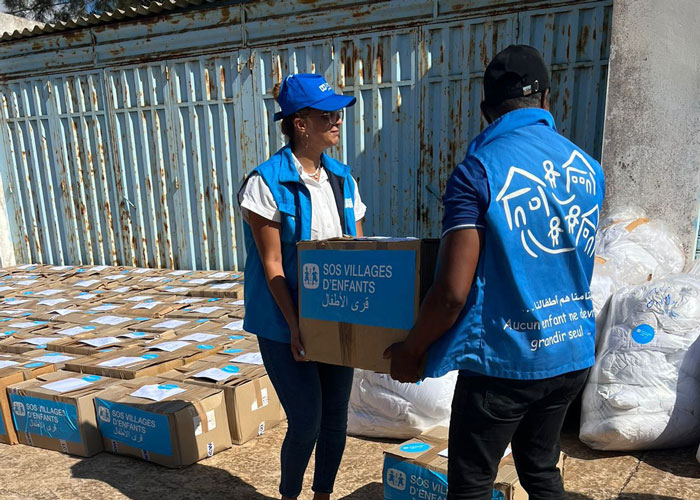 To medarbeidere fra SOS-barnebyer med blå vester med logo på, bære er eske med nødhjelpsartikler mellom seg. I bakgrunnen står mange kasser. Foto: SOS-barnebyer