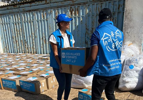 To medarbeidere fra SOS-barnebyer med blå vester med logo på, bære er eske med nødhjelpsartikler mellom seg. I bakgrunnen står mange kasser. Foto: SOS-barnebyer