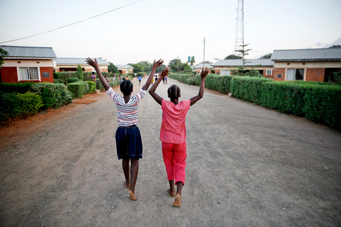 To jenter fra SOS-barnebyen Livingstone går på veien mellom husene og strekker armene i været. Foto: Paul Audestad
