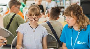 En kvinnelig SOS-barnebyers medarbeider med blå t-skjorte med logo og mørkeblondt har, sitter ved et bord sammen med ei jente med lys lilla t-skjorte, blondt hår  hestehale og briller. Foto: Borysova Kseniia