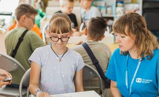 En kvinnelig SOS-barnebyers medarbeider med blå t-skjorte med logo og mørkeblondt har, sitter ved et bord sammen med ei jente med lys lilla t-skjorte, blondt hår  hestehale og briller. Foto: Borysova Kseniia