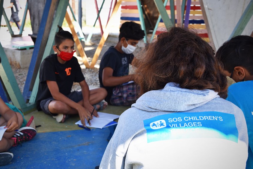SOS-ansatt med ryggen til underviser barn i en flyktningleir. De sitter inne i et telt. 