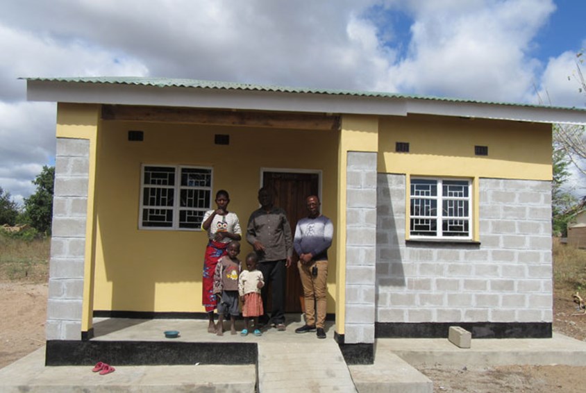 En familie med to av barna sine står utenfor huset sammen med en medarbeider fra SOS-barnebyer. Huset er lagd av mursteinsblokker, har vinduer med sprosser, bølgeblikktak og deler av huset er malt gult. Foto: SOS-barnebyer