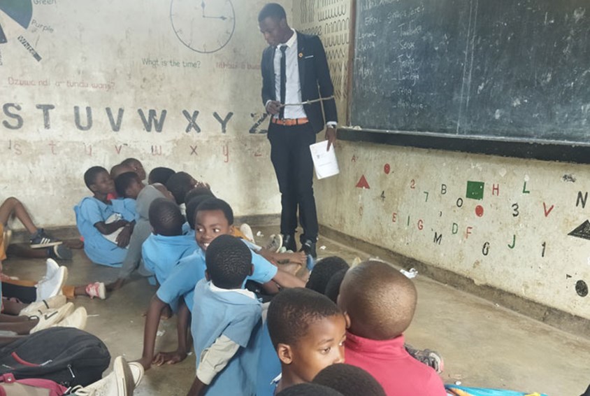 Yohane, i dress, hvit skjorte og slips, står foran tavla og underviser. Foran ham sitter elevene på gulvet. Foto: SOS-barnebyer