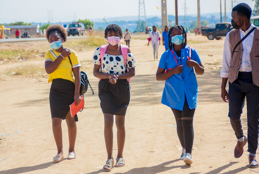 Fire ungdommer i fargerike klær og med munnbind går til skolen langs veien og prater sammen. Foto: André Canduco
