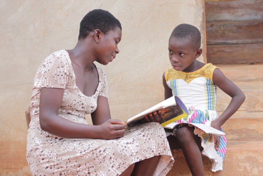 Abena, med mørkt kort hår og rosa kjole, hjelper lillesøster med leksene. Foto: Nana Sampson