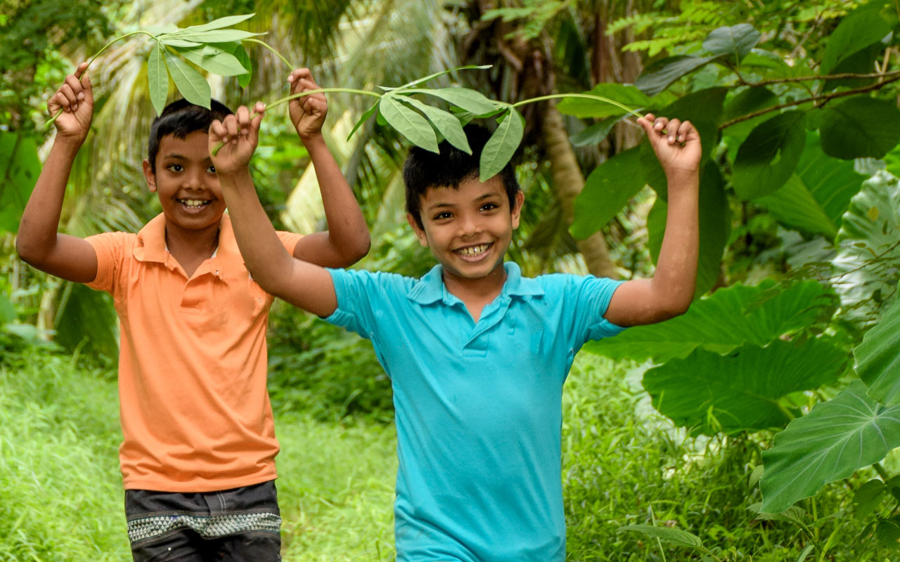 To gutter fra Sri Lanka, den ene i oransje t-skjorte og den andre i turkis, strekker hendene i været med et stort palmeblad over hodet. Begge smiler. Foto: Pearl Sandhu