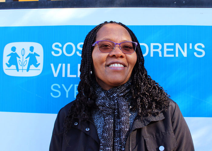  Teresa Ngigi, psykolog ved SOS-barnebyer, har langt, mørkt hår og briller, hun står foran et skilt med SOS-barnebyers logo. Foto: Lur Katt Web