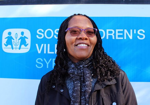  Teresa Ngigi, psykolog ved SOS-barnebyer, har langt, mørkt hår og briller, hun står foran et skilt med SOS-barnebyers logo. Foto: Lur Katt Web