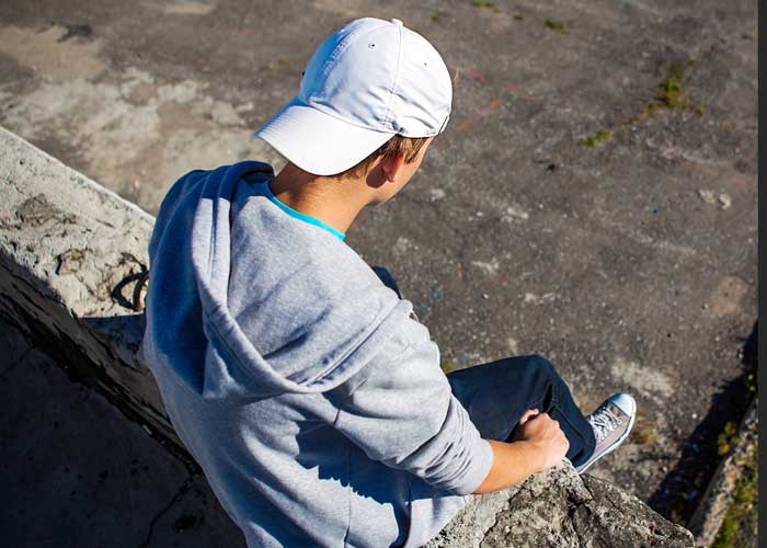 Gutt med grå collegegenser, hvit cap og jeans sitter på en mur. Bildet er tatt bakfra. Foto: SOS-barnebyer