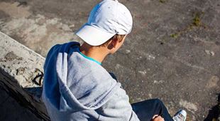 Gutt med grå collegegenser, hvit cap og jeans sitter på en mur. Bildet er tatt bakfra. Foto: SOS-barnebyer
