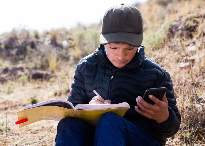 En gutt fra SOS-barnebyers familieprogram i Peru sitter ute og følger undervisningen via mobilen. Han har blå boblejakke, blå bukse og caps på seg, og skriver konsentrert i ei bok. Foto: Alejandra Kaiser
