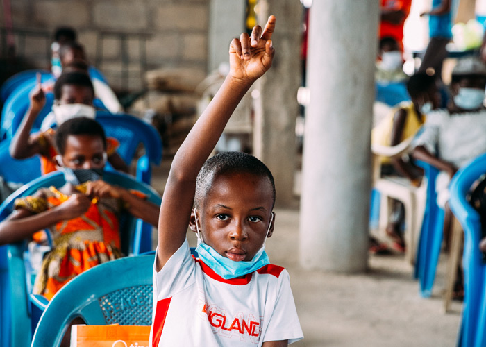 Gutt i hvit og rød t-skjorte, som er en del av SOS-barnebyers familieprogram i Ghana, sitter ved pulten sin og rekker opp hånda. Foto: Alea Horst