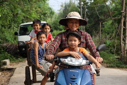 En mor med hatt kjører moped, med et barn foran i oransje t-skjorte og tre barn bak. Fra Vietnam.