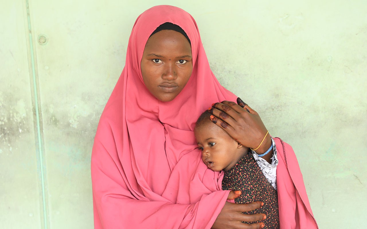 Mor med rosa hijab sitter inntil en grønn vegg og holder babyen sin tett inntil seg. Foto: SOS-barnebyer