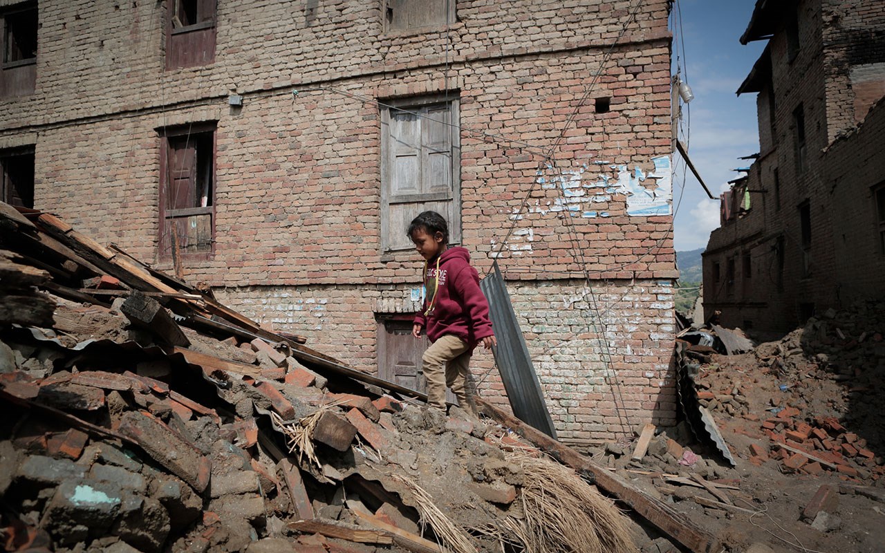 Jente med mørkt hår, hettegenser og bukse går i ruinene etter et hus som har rast sammen. Foto Zishaan Akbar Latif