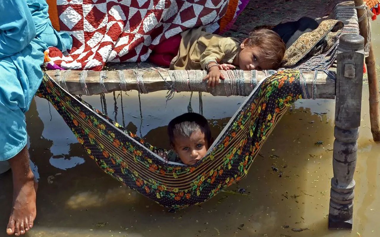 Ett barn ligger blant noen tepper i en provisorisk beskyttelse som står på påler, ett barn ligger og titter ut av ei hengekøye som henger under gulvet. Det er vann på alle kanter. Foto Naseebullah Jamali