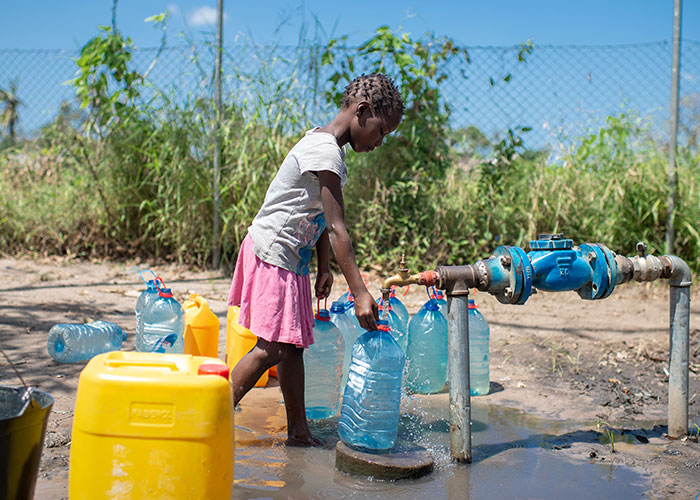 Jente fra Mosambik med fletter, hvit t-skjorte og rosa skjørt fyller vann på ei plastkanne, fra en vannpost. Foto: Cornel van Heerden