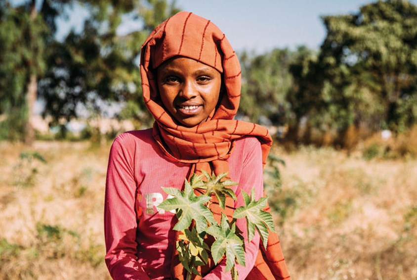 Jente står i åkeren og holder en grønn vekst i hånda. Hun har på seg oransje hijab og rød genser. Foto: Alea Horst