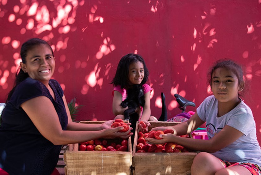 Moren, Slivia, som holder den lille valpen, og søsteren sitter rundt to kasser med røde epler. Veggen bak dem er rosa. Foto Hairo Caba/Monica Garcia