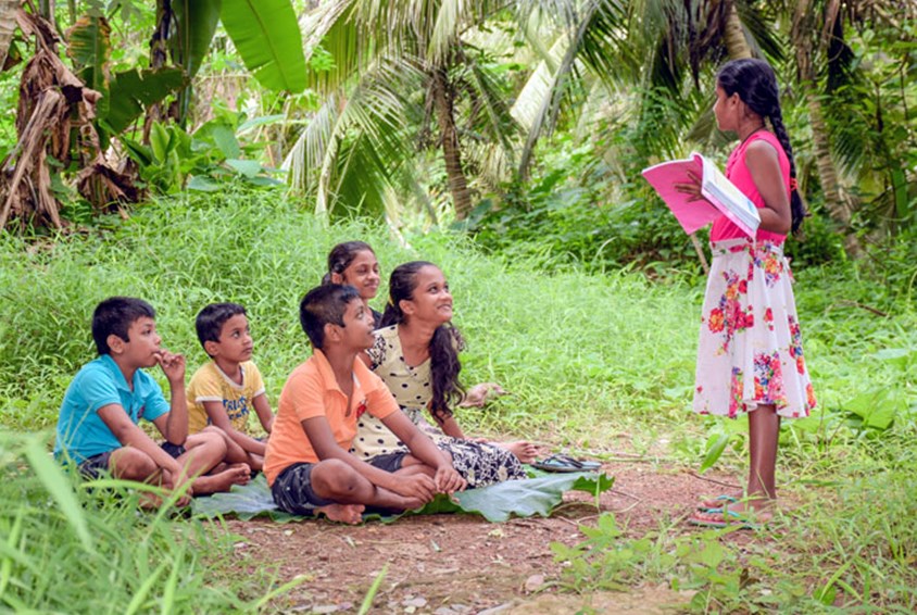 Samith, brødrene og noen andre barn sitter på gresset. Ei jente i rosa kjole står foran dem og leser i ei bok. Foto: Pearl Sandhu