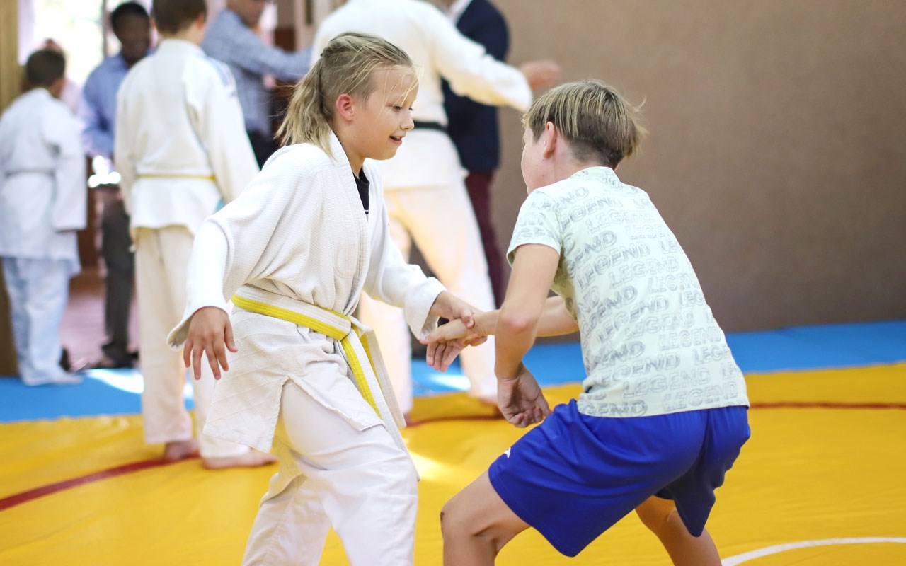 En jente i hvit jiu-jitsu klær med gult belte, har tatt tak i en gutt. Han har grønn t-skjorte og blå shorts. Foto: SOS-barnebyer