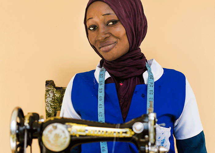 Maimuna sitter ved symaskinen og smiler stolt. Hun har et svart hodeplagg, hvit og blå skjorte. Foto Mona van den Berg
