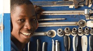Ung gutt fra Gambia i blå kjeledress står ved en vegg full av verktøy og smiler. Foto: Claire Ladavicius