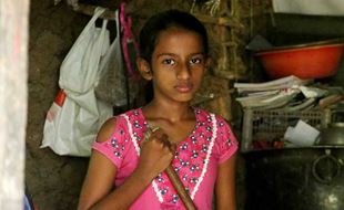 Alvorlig jente fra Sri Lanka med mørkt langt hår i hestehale og rosa kjole, står på kjøkkenet og holder i en kost. Foto: Pearl Sandhu
