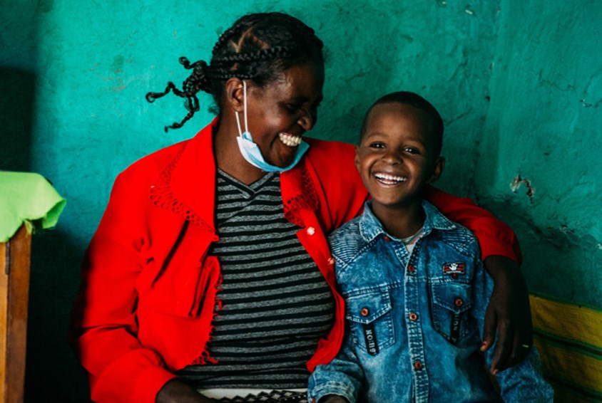 Mor i rød jakke, stripete skjorte og med munnbindet på haken holder et godt tak rundt gutten sin som har på seg jeansjakke. Begge smiler stort. Foto: Alea Horst. Fra SOS-barnebyers familieprogram i Etiopia. 