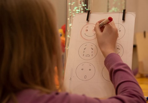 Jente med langt lyst hår og lys lilla genser tegner runde ansikter med forskjellige uttrykk, et med smil, et med tårer og et som ser forundret ut. Foto: Katerina Ilievska