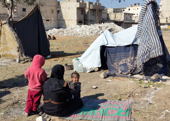 En mor og to barn sitter på bakken i et område hvor det er satt opp provisoriske telt. Foto Mahmoud Shabarek