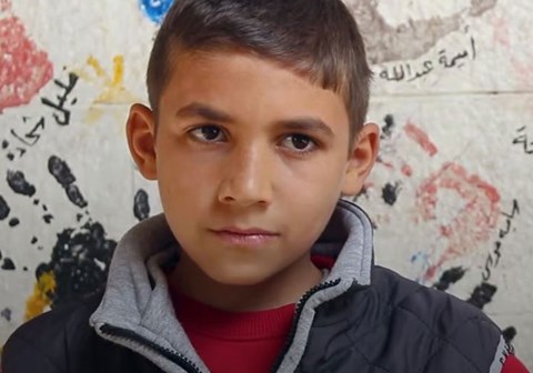 Gutt med kort mørkt hår, blå vest og rød genser står foran en vegg malt med håndavtrykk. Foto: SOS-barnebyer
