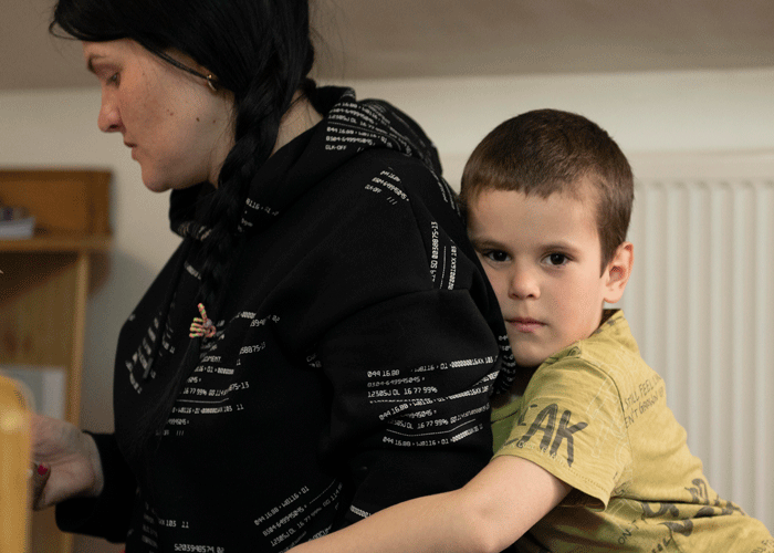 Gutt i gulgrønn t-skjorte lener seg inntil morens rygg og ser alvorlig inn i kamerat. Foto: Katerina Ilievska