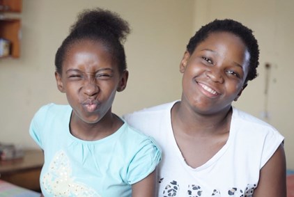 To søstre fra Sør-Afrika. Den ene som har oppsatt hår og lyseblå t-skjorte gjør en morsom grimase den andre har kort hår og hvit s-skjorte, hun smiler. 
