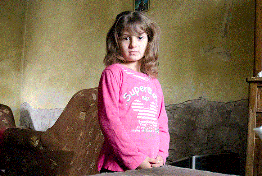 Jente i rosa genser og mørkt halvlangt hår står i et rom med slitte murvegger. Foto: SOS-barnebyer