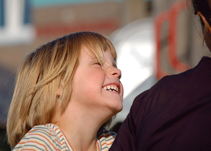 Jente i profil, med lyst hår og stripete genser, sitter inntil moren sin og ler. Foto: Marko Mägi