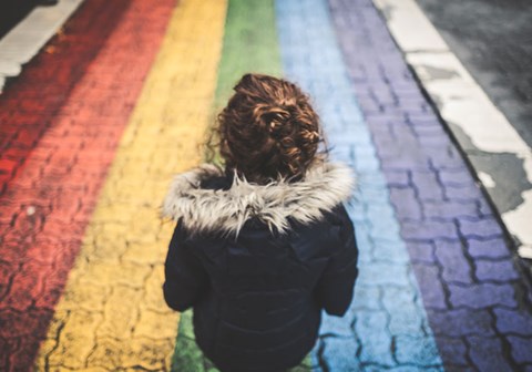 Jente med krøllete hår og blå jakke med pelskrage står med ryggen til i ei gate farget i regnbuens farger. Foto: Cory Woodward on Unsplash